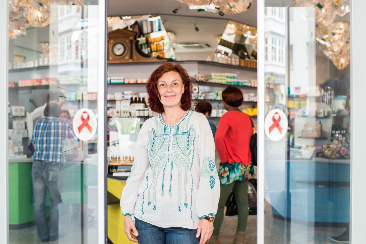 Mag. pharm. Karin Simonitsch steht zwischen den Schiebetüren im Eingang der Marien Apotheke. An den Schiebetüren sind links und rechts Sticker mit dem Red Ribbon für HIV-Awareness angebracht.