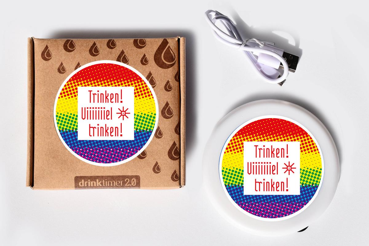 Der intelligente Untersetzer mit Trinkerinnerung, der Drink Timer, in einer Schachtel und daneben offen mit Ladekabel. Der Drink Timer ist von der Marien Apotheke Wien individualisiert und der Spruch auf dem regenbogenbunten Sticker lautet 