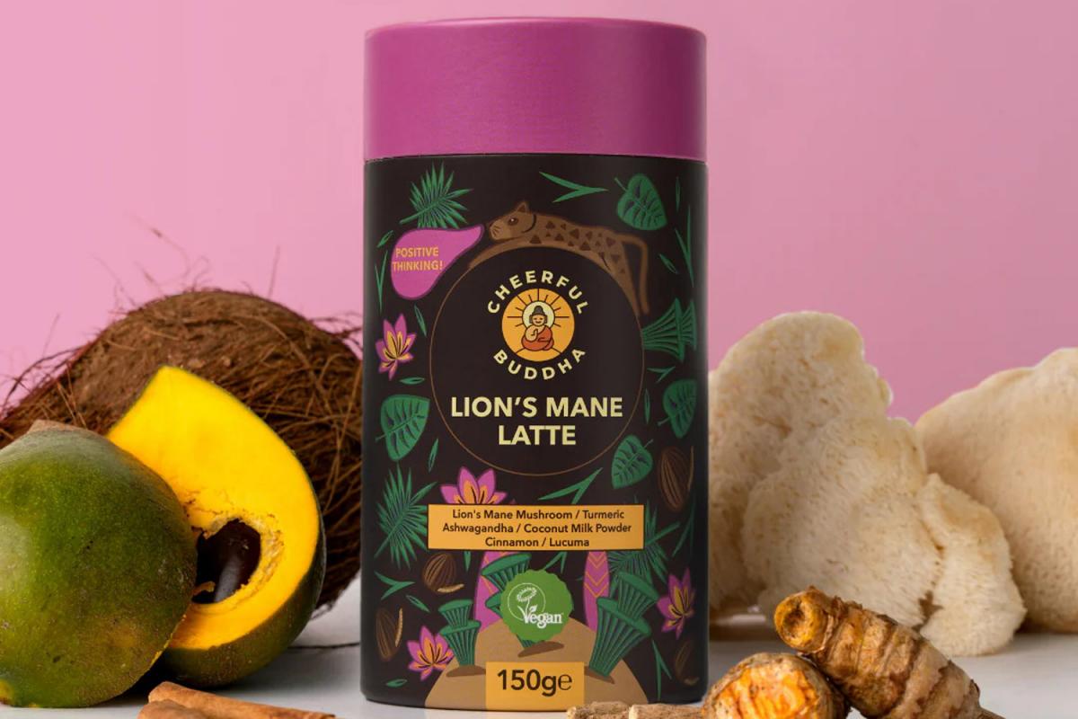 Lion's Mane Latte von Cheerful Buddha in der Verpackung. Links daneben liegt eine Mango und eine Kokosnuss, rechts daneben der Lions Mane Pilz und Kurkuma-Wurzel.